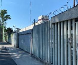 Calle junto a valla metálica con alambre de púas bajo un cielo despejado.