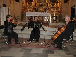 Trío de cuerdas tocando en una iglesia, con violín, viola y violonchelo frente a un altar.