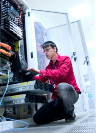 Techniker in rotem Hemd arbeitet in einem Serverraum mit einem Laptop an Netzwerkgeräten.