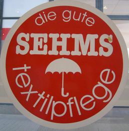 Rotes Schild mit Text "SEHMS Textilpflege" und einem Regenschirm-Symbol in der Mitte.