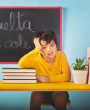 Niño en clase con expresión pensativa, pizarra dice "Vuelta al cole", libros y planta en mesa.