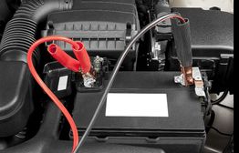Car battery with red and black jumper cables attached in engine compartment.