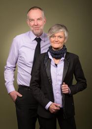 Ein Mann und eine Frau in formeller Kleidung lächeln vor einem neutralen Hintergrund.