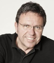 Ein lächelnder Mann mit Brille und dunklem Shirt vor einem neutralen Hintergrund.