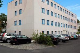 Mehrstöckiges Gebäude mit geparkten Autos davor, klare blaue Fenster, sonniger Himmel.