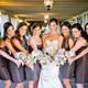 A bride and bridesmaids in brown dresses smile, holding bouquets in a decorated venue.