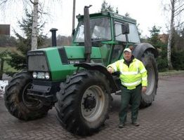 Eine Person in Warnweste steht neben einem großen grünen Traktor auf einem gepflasterten Weg.