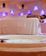 Persona en bañera y otra en mesa de masaje, ambiente con luces violetas.