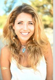 Smiling woman with wavy hair in a white top, wearing earrings and a blue pendant necklace.