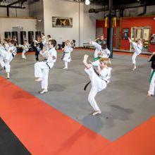 Students practicing martial arts kicks in a spacious dojo with red and gray flooring.