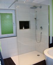 Moderne Dusche mit Glaswänden, weiß gefliest, neben einem Fenster mit grünem Sichtschutz.
