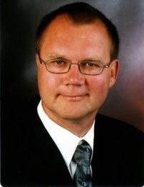 Ein Mann mit Brille und Anzug lächelt leicht in die Kamera.