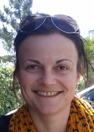 Frau mit Sonnenbrille im Haar, lächelnd, trägt einen gelben Schal, Hintergrund mit Bäumen.