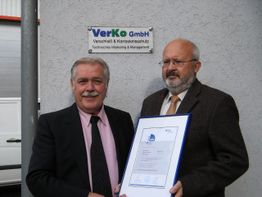 Zwei Männer stehen vor einem Firmenlogo und halten ein Zertifikat in Händen.