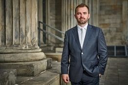 Ein Mann im Anzug steht vor alten Steinsäulen und blickt in die Kamera.