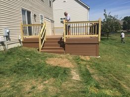Man standing on new wooden deck with railing by a house, surrounded by grassy yard.