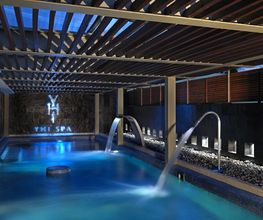 Piscina interior iluminada en azul, con fuentes de agua y techo de madera en un spa moderno.