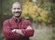 Man in a red plaid shirt smiling with crossed arms, standing outside with autumn trees behind.