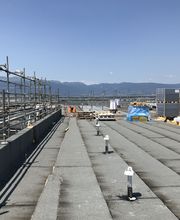 Baustelle auf einem Flachdach mit Gerüsten, Bergen im Hintergrund und blauem Himmel.