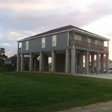 Elevated house on tall stilts with a grassy yard, set under a cloudy sky.