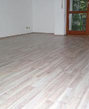 Leerer Raum mit hellem Laminatboden, weißer Wand und großem Fenster mit Blick ins Grüne.