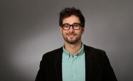 Ein Mann mit Brille lächelt, trägt ein türkisfarbenes Hemd und dunkles Sakko vor grauem Hintergrund.