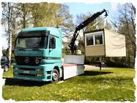 Ein Lkw mit Kran hebt ein kleines Gebäude auf einer Wiese an.