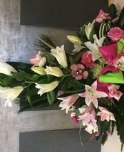 Arreglo floral en una cruz: lirios blancos y rosados, con hojas verdes sobre fondo gris.