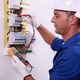 Electrician in blue uniform and helmet testing wires with a multimeter.