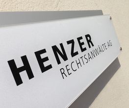 Hinweisschild an einer Wand mit der Aufschrift "HENZER RECHTSANWÄLTE AG".