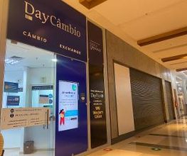 Fachada de casa de câmbio em shopping, com letreiros iluminados e portas de metal fechadas.