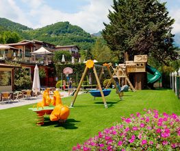 Spielplatz mit Schaukeln, Rutsche und Wippe auf Rasen, umliegende Berge und Gebäude im Hintergrund.
