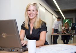 Eine Frau lächelt am Laptop; im Hintergrund stehen zwei Personen an einem Tisch im Büro.
