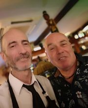 Dos hombres sonríen en un bar bien iluminado. Uno viste camisa formal, el otro camisa floral.