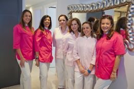 Seis mujeres con batas rosadas y blancas posan sonriendo junto a un espejo decorativo.