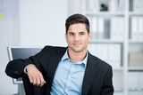 Ein lächelnder Mann in Anzug sitzt entspannt im Büro und schaut in die Kamera.