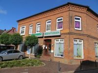 Backsteingebäude mit Schildern, davor Bäume und ein geparktes Auto unter blauem Himmel.