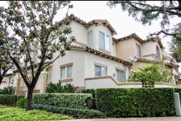 Two-story beige house with tile roof, bushes, and trees, viewed from the corner of a street.