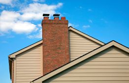Brick chimney on a beige house roof against a clear blue sky with a few clouds.