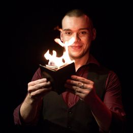 Ein Mann hält ein brennendes Buch und lächelt in die Kamera, dunkler Hintergrund.