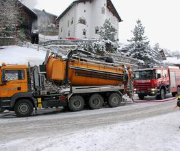 LKW und Feuerwehrfahrzeug im Schnee vor Häusern; Feuerwehrleute in Warnwesten sind zu sehen.