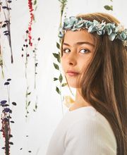 Niña con corona de flores azules, rodeada de ramas y plantas colgantes en fondo claro.