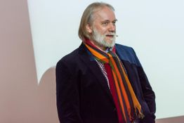 Ein Mann mit buntem Schal hält einen Vortrag vor einer hellen Wand.