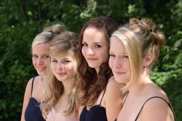Vier junge Frauen in eleganten Kleidern draußen, im Hintergrund grüne Blätter.