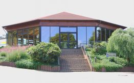 Achteckiges Gebäude mit Glasfassade, Treppe und umgeben von grüner Landschaft und Sträuchern.