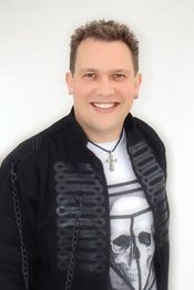 Ein lächelnder Mann mit Kette, schwarzer Jacke und T-Shirt mit Totenkopf-Design auf weißem Hintergrund.