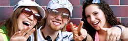Drei junge Erwachsene lachen und zeigen das Peace-Zeichen vor einer Kachelwand.