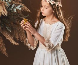 Niña con corona de flores y vestido blanco, observando un ramo de hojas secas y flores.