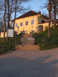 Gelbes Haus auf einem Hügel, von einer Treppe umgeben, Bäume im Vordergrund, bei Sonnenuntergang.
