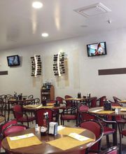 Ristorante con tavoli apparecchiati, pareti chiare, TV e bottiglie esposte. Ambiente accogliente.